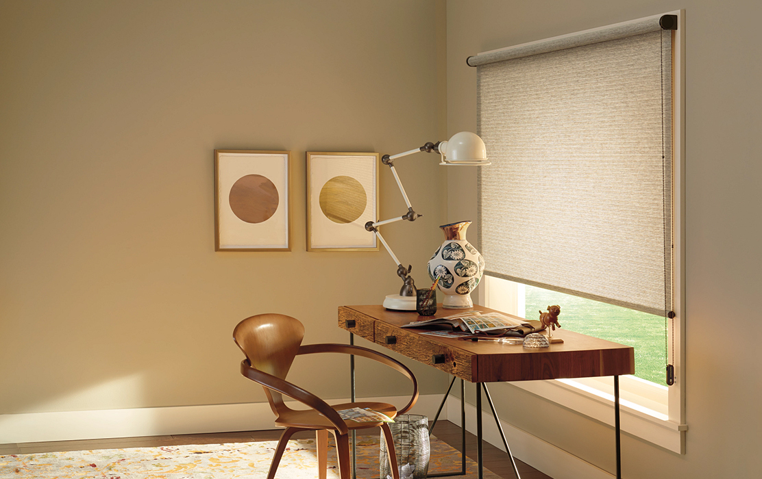 A stylish home office with a modern desk, chair, and a window adorned with elegant designer roller shades, providing privacy and diffused light.
